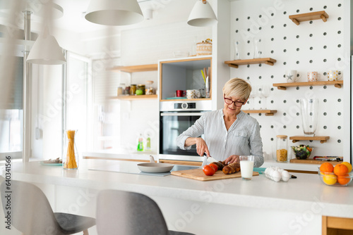 Photo of concentrated Caucasian senior woman making healthy snacks for her family in modern minimalist kitchen.