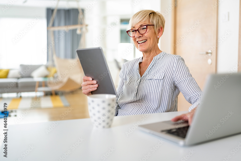 Satisfied happy senior woman reading on tablet while texting on laptop and sitting at dining table. Satisfied happy senior woman reading on tablet while texting on laptop and sitting at dining table.