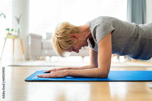 Concentrated determined middle aged blonde woman doing plank on yoga mat at home.