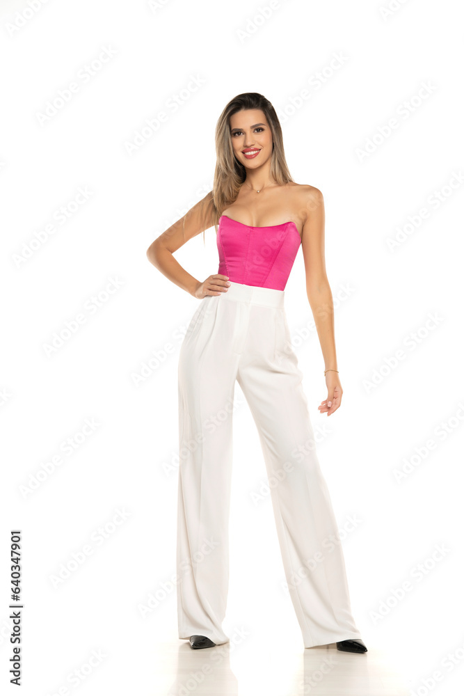 young modern smiling woman in white pants and pink corset posing on white background