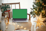 Laptop with empty screen and stylish christmas gifts on modern chair on background of festive decorated tree, golden lights and star in room. Christmas shopping online and sales. Laptop mock up