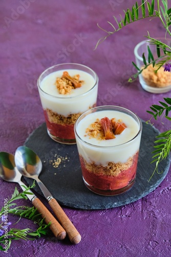 Portioned dessert in a glass with rhubarb jam, shortbread crumble and custard on a purple concrete background. Rhubarb recipes. British cuisine.