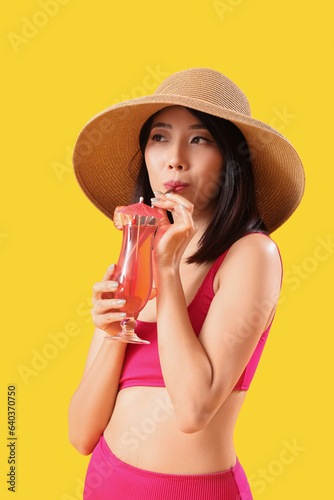 Beautiful Asian woman in swimsuit and with cocktail on yellow background