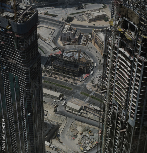 Aerial view looking down onto skyscrapers under construction in Dubai. The unfinished buildings soar high. The background behind them is a mix of ongoing construction and finished projects.