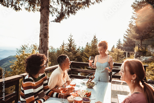 Young and diverse group of female friends enjoying a glass of wine and dinner on a balcony in a cabin house with a scenic view of the mountain and forest below photo