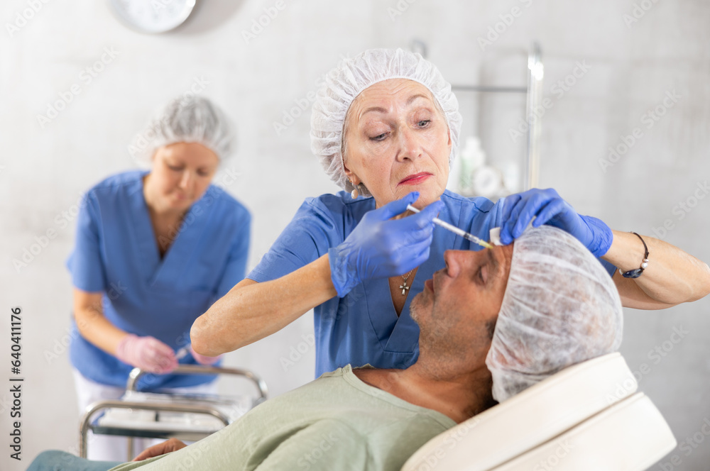 Obraz premium Team of cosmetologists provide anti-aging injection services to male patient. Elderly female doctor performs wrinkle smoothing procedure, gives injection of botulinum toxin to man patient in forehead