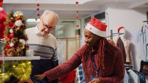 Helpful retail assistant wearing santa hat during festive holiday season helping old man with best fitting blazer. Employee assisting customer in Christmas decorated clothing store