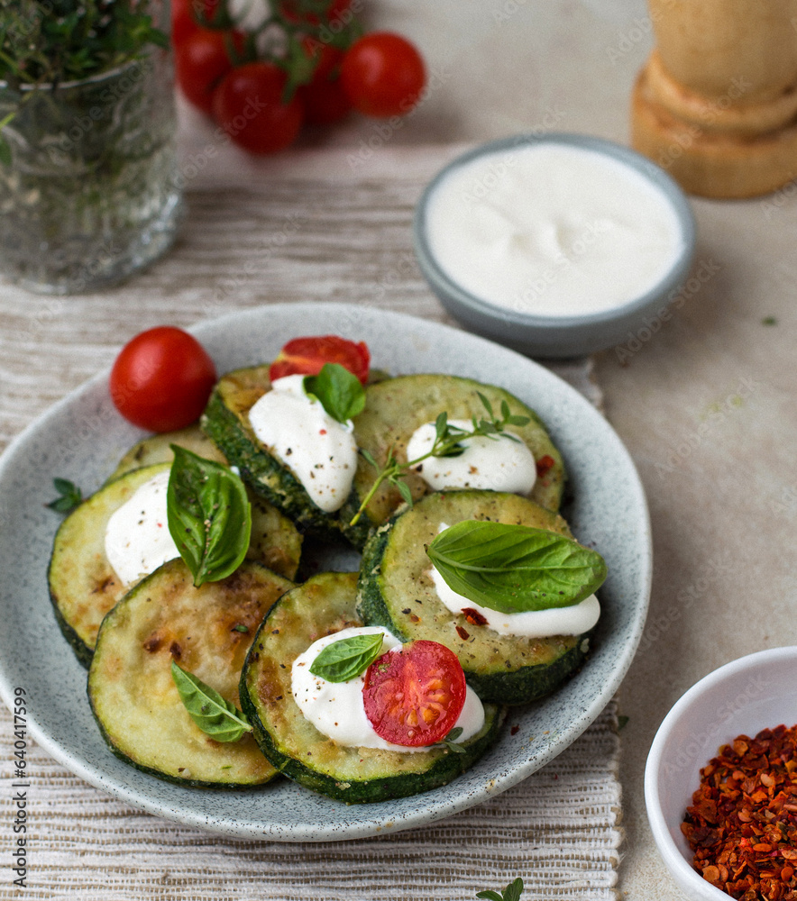 Zucchini fried on a plate with basil and cherry tomatoes, sour cream sauce and spices in bowls, cherry sprigs, herbs, pepper mill.