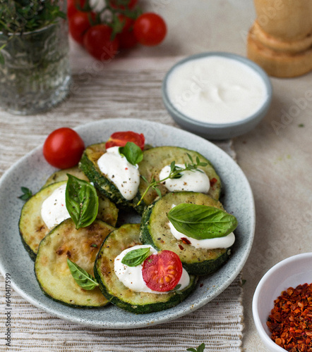 Zucchini fried on a plate with basil and cherry tomatoes, sour cream sauce and spices in bowls, cherry sprigs, herbs, pepper mill.