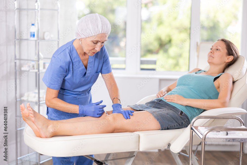 Obraz premium Elderly female doctor in medical uniform makes injection in leg of adult female patient in medical room