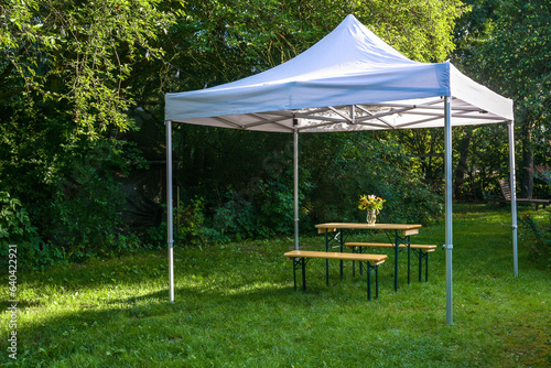 Fototapeta Naklejka Na Ścianę i Meble -  White gazebo or pavilion with a wooden table, benches and a flower bouquet on the meadow in a natural garden, idyllic summer place gives protection from sun and rain, copy space