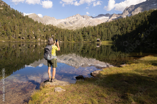 Gray-haired man with backpack photographing beautiful landscape with mountain lake and reflection, Austria