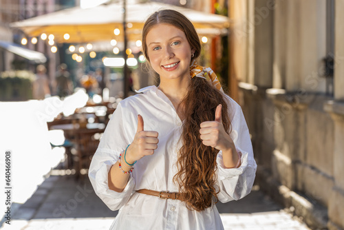 Like. Happy redhead teenager child girl looking approvingly at camera showing thumbs up like sign positive something good positive feedback. Woman standing on city street. Town lifestyles outdoors