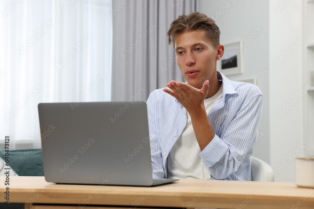 Happy young man having video chat via laptop and blowing kiss at wooden table indoors. Long-distance relationship Happy young man having video chat via laptop and blowing kiss at wooden table indoors. Long-distance relationship
