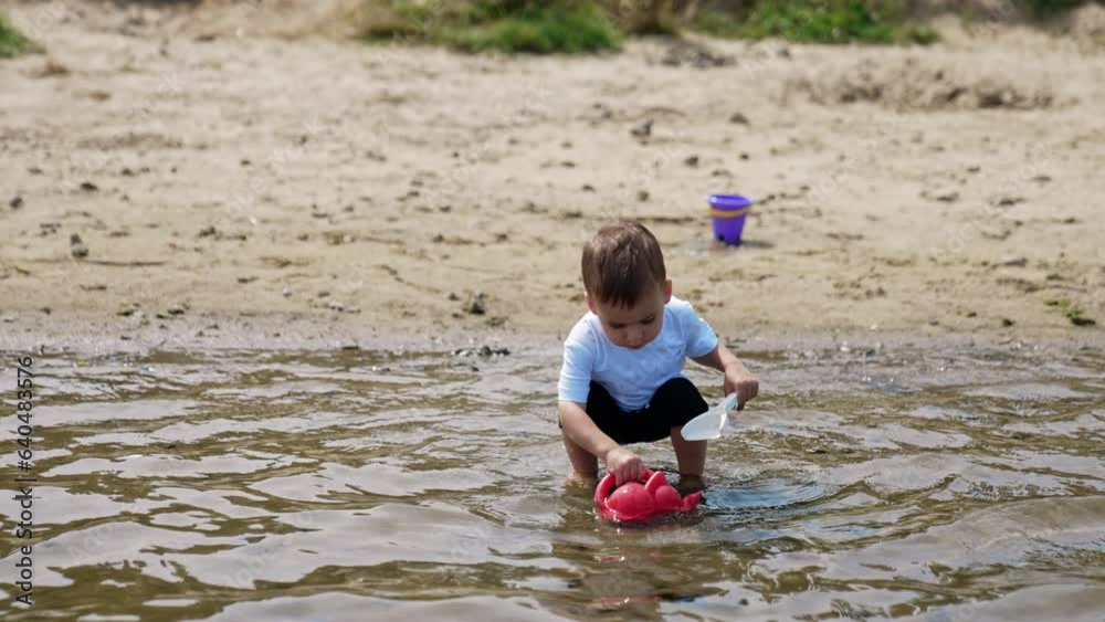 Charming baby boy picks some water from the river. Kid walks by the water holding a watering can and a shovel. Charming baby boy picks some water from the river. Kid walks by the water holding a watering can and a shovel.