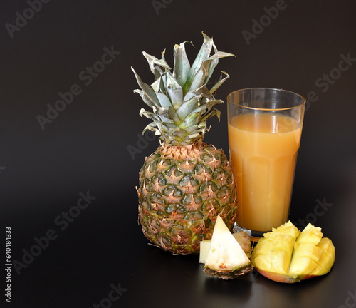 A tall glass of fresh fruit juice on a black background, next to pieces of ripe pineapple and mango.