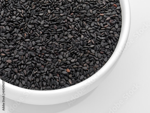 Close-up of black sesame seeds in a white bowl on a white background, top view, flat lay, cup partially. Horizontal.