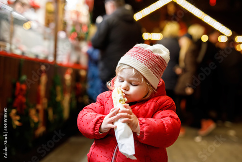 Little baby girl, cute child eating bananas covered with chocolate, marshmellows and colorful sprinkles near sweet stand with gingerbread and nuts. Happy toddler on Christmas market in Germany.