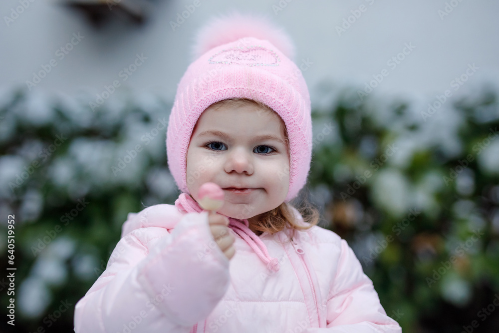 Portrait of little toddler girl walking outdoors in winter. Cute toddler eating sweet lollypop candy. Child having fun on cold snow day. Wearing warm baby pink clothes and hat with bobbles. Portrait of little toddler girl walking outdoors in winter. Cute toddler eating sweet lollypop candy. Child having fun on cold snow day. Wearing warm baby pink clothes and hat with bobbles.