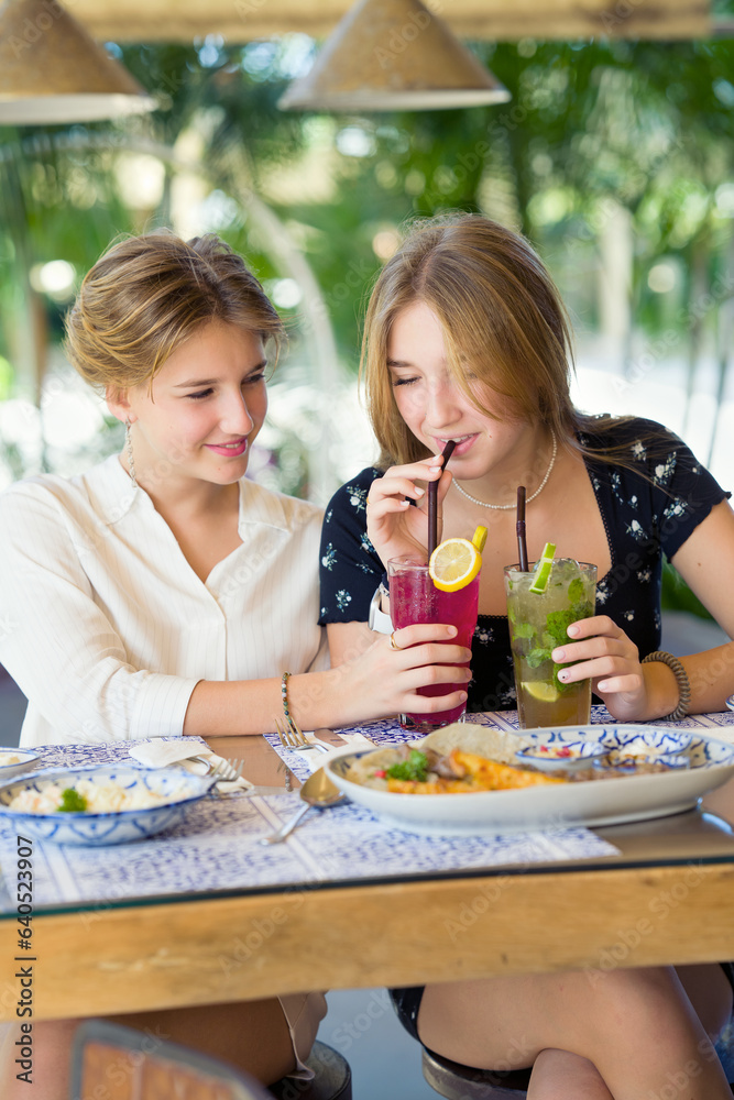 Two happy teenage young girls met each other in the cafe drinking fruit cocktails. Summer warm day on vacations.
