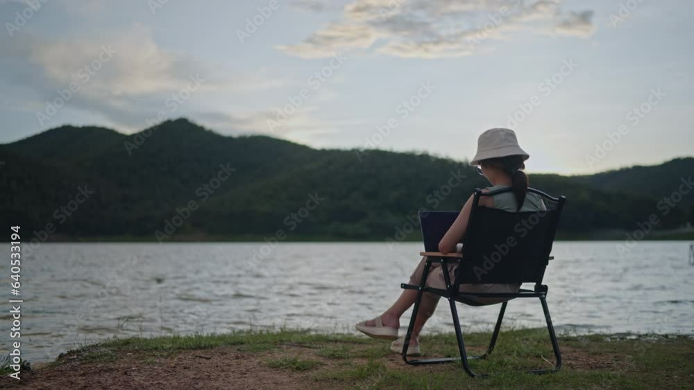 Woman working on laptop by the lake and mountain in evening with golden sunset. Travel to nature place.