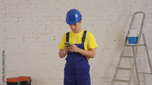 Medium shot of a young construction worker standing in the room, holding a smartphone, waiting for the answer. The man looks intemperate and concerned. photo