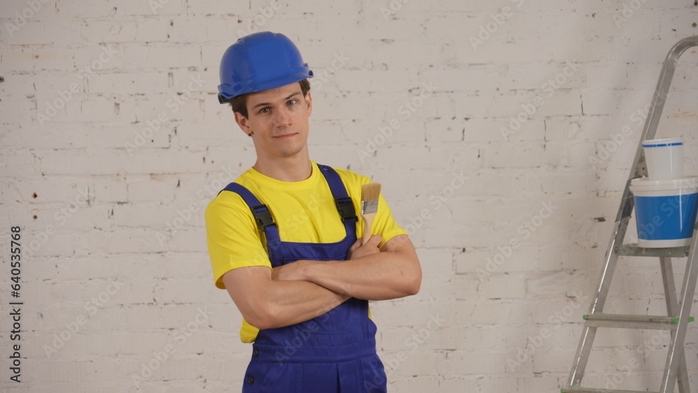 Medium shot of a young construction worker standing in the room under renovation with a brush in his hand, crossing his arms with confidence. Medium shot of a young construction worker standing in the room under renovation with a brush in his hand, crossing his arms with confidence.