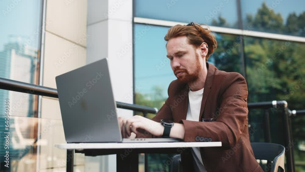 Emotional red-haired businessman typing on a laptop and working remotely outdoors. A busy male worker in a formal suit solves business problems against the backdrop of a building and writes an email. Emotional red-haired businessman typing on a laptop and working remotely outdoors. A busy male worker in a formal suit solves business problems against the backdrop of a building and writes an email.