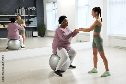 African American plump woman training on fitness ball together with instructor in health club