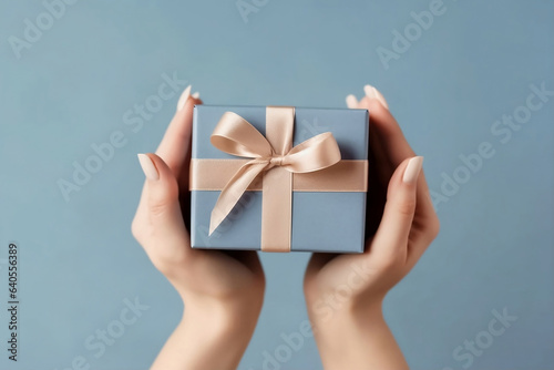 Stampa su tela Female hands with natural manicure holding blue gift box with light golden ribbon on trendy beige background