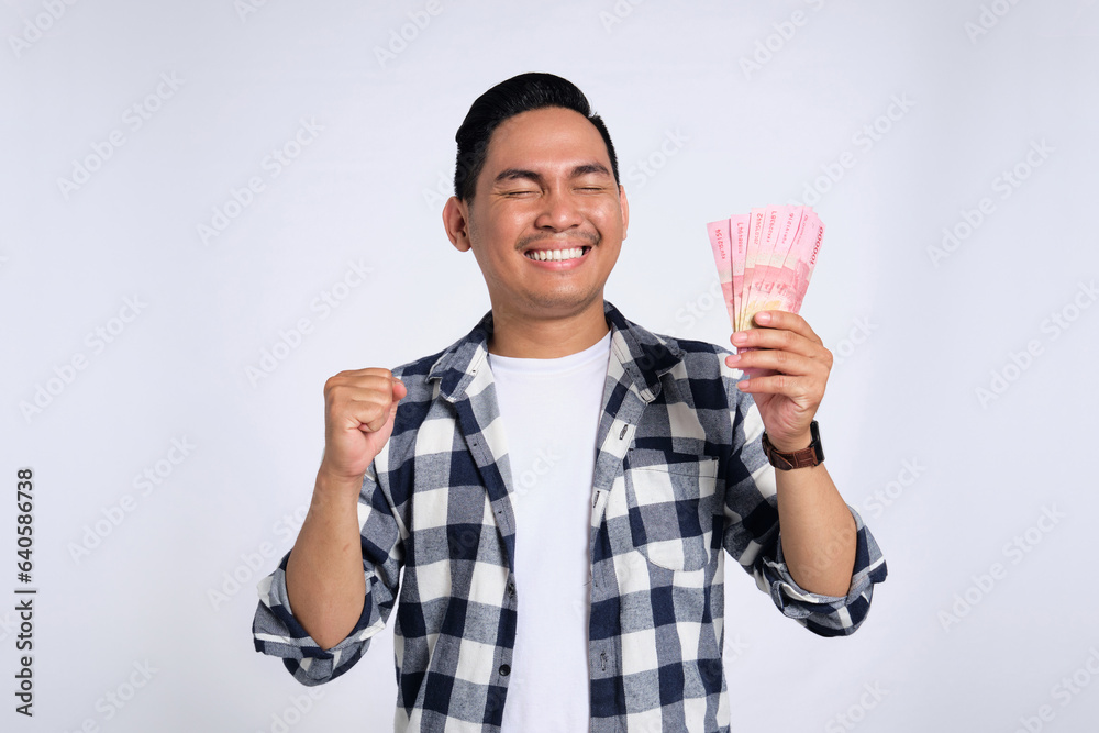 Excited young Asian man in casual shirt holding money and clenching fist, celebrating financial success isolated on white background. Profit and wealth concept Excited young Asian man in casual shirt holding money and clenching fist, celebrating financial success isolated on white background. Profit and wealth concept
