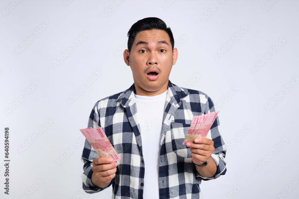 Surprised young Asian man in casual shirt holding money and looking at camera with confused expression isolated on white background Surprised young Asian man in casual shirt holding money and looking at camera with confused expression isolated on white background