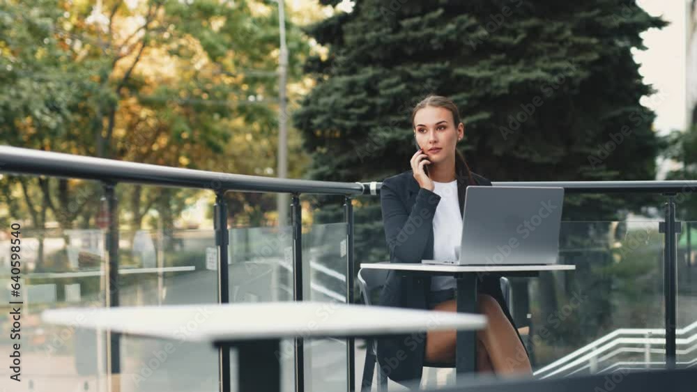 Freelancer with a laptop on the outdoor terrace of a cafe. A brunette woman uses a laptop outdoors, dressed in a classic suit. A woman is using a laptop for remote work and talking on the phone. Freelancer with a laptop on the outdoor terrace of a cafe. A brunette woman uses a laptop outdoors, dressed in a classic suit. A woman is using a laptop for remote work and talking on the phone.