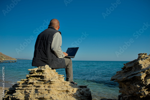 A man with a laptop sits on the seashore and works. Remote remote work from anywhere in the world