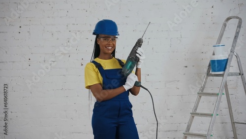 Medium shot of a dark-skinned young female worker standing in the room under renovation, looking around, raising the drill, perforator and holding it like a gun.