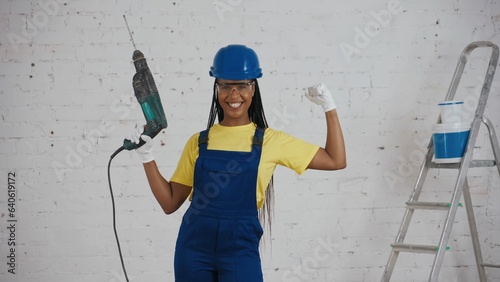 Medium shot of a dark-skinned young female worker standing in the room under renovation, looking around, raising the drill, perforator and showing her muscles.