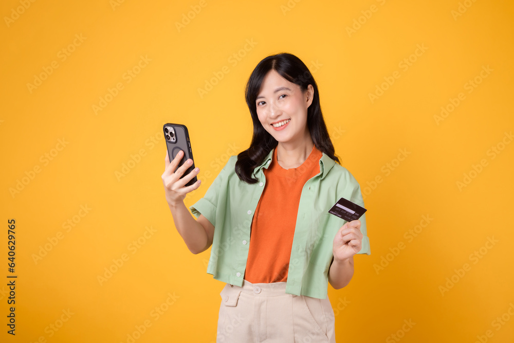 the convenience of mobile shopping with an enchanting young Asian woman in her 30s, wearing orange shirt and green jumper, using smartphone while holding credit card on yellow studio background. the convenience of mobile shopping with an enchanting young Asian woman in her 30s, wearing orange shirt and green jumper, using smartphone while holding credit card on yellow studio background.