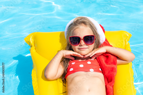 Cute happy little girl in Santa Claus hat, sunglasses and red swimsuit lying on inflatable mattress in swimming pool on tropical vacation. Christmas in warm countries. Advertising for tour operators
