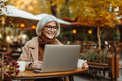 Aged woman uses her laptop in autumn park. Digital era, tech-savvy senior and modern technology