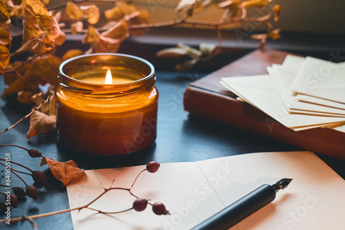 Cozy autumn still life fall leaves, sheets of paper, fountain pen and burning candle on the windowsill. Autumn home decor. Cozy fall mood.