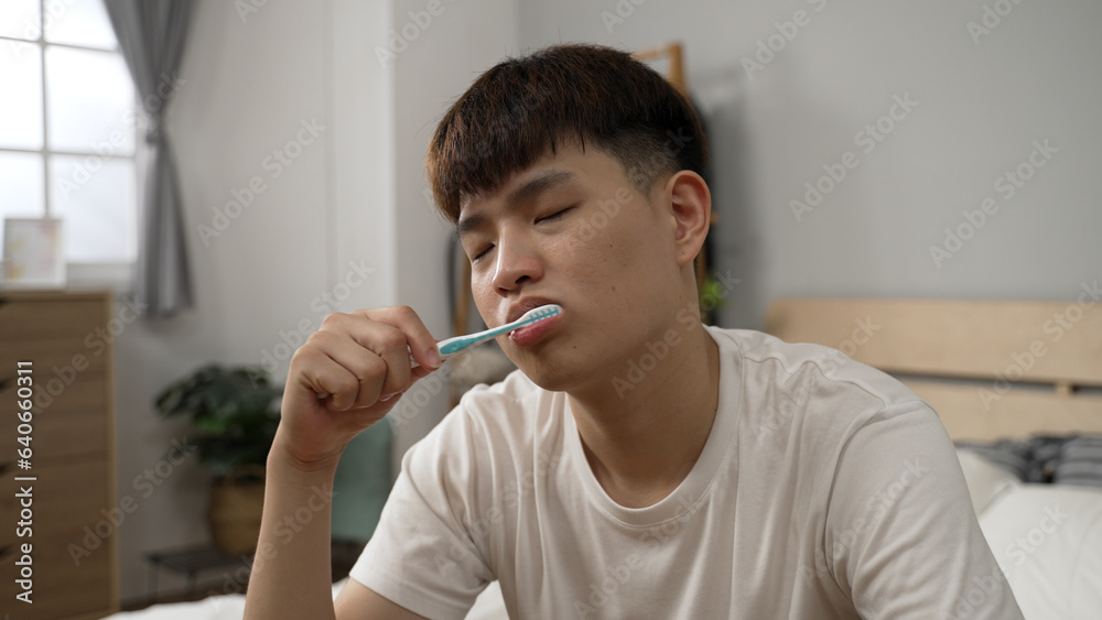 closeup shot of an asian man in casual wear is brushing teeth with fatigue after getting up on Monday morning in the bedroom at home. closeup shot of an asian man in casual wear is brushing teeth with fatigue after getting up on Monday morning in the bedroom at home.