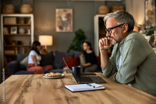 A senior man working on a laptop at home, while his family sitting on a couch.