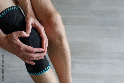 Black knee support brace on a man leg. Guy in an orthosis at home. Orthopedic anatomic braces for knee fixation, injuries and pain. Knee Joint Bandage Sleeve. Elastic Sports.