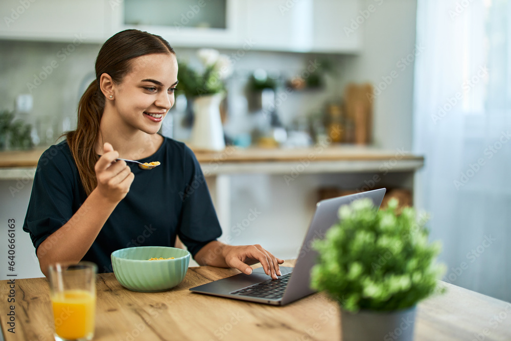 A smiling female freelancer eating cereal for breakfast while working on a laptop.