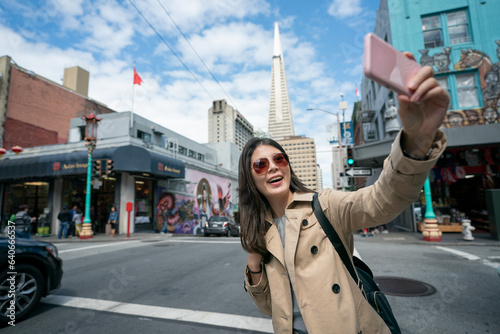 happy asian Taiwanese woman traveler holding phone and having fun taking selfie photo with landmark Transamerica Pyramid at street corner in san Francisco's china town