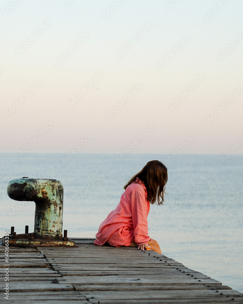 A girl, in a long pink shirt, greets the morning on a pier in the ocean, thinking about life.