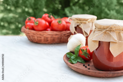 A sauce of fresh red tomatoes in a glass jar on a wooden plate. Sprig of fresh tomatoes, garlic, pepper and basil on a white table. A can of homemade ketchup with blurred green background. Copy space.