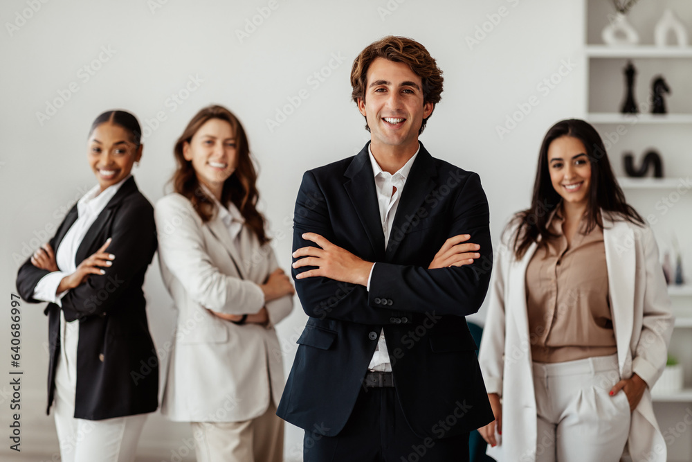 Positive businessman leader standing with folded arms in front of his multiracial female employees, posing in office Positive businessman leader standing with folded arms in front of his multiracial female employees, posing in office