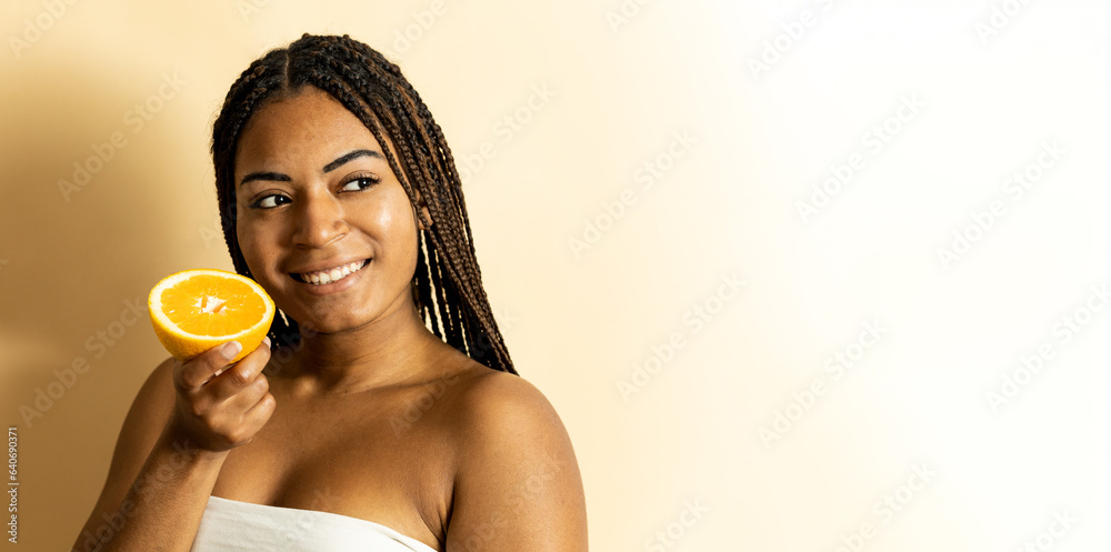 A cute young African woman with braided hair poses against an orange background. The woman holds half an orange with one hand while looking sideways.Nutrition with vitamin C for skin health.Copyspace A cute young African woman with braided hair poses against an orange background. The woman holds half an orange with one hand while looking sideways.Nutrition with vitamin C for skin health.Copyspace