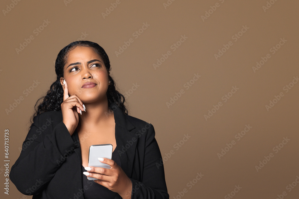 Thoughtful brazilian plus size woman using cellphone and looking at copy space on brown background, place for your ad Thoughtful brazilian plus size woman using cellphone and looking at copy space on brown background, place for your ad